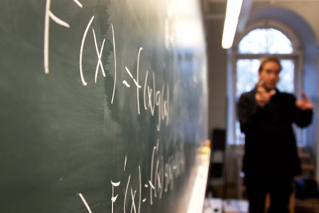 A greenish blackboard with white text. A person teaching is seen, blurry, in the background. Photo.