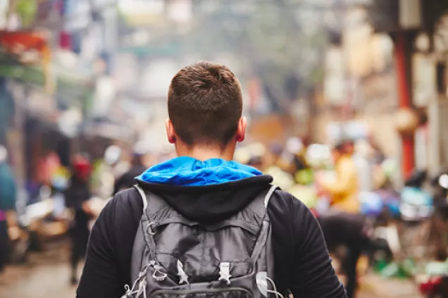 Photograph of a traveller from behind. He is wearing a black jacket and a backpack, looking over a colourful city. 