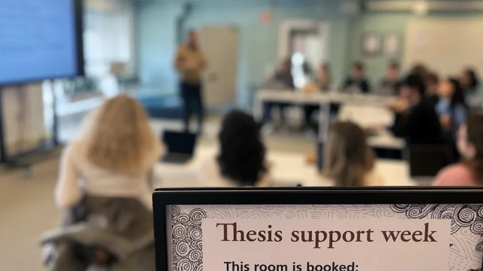 In the foreground a sign which reads "thesis support week". In the background a lecture with sutdents. Photo.
