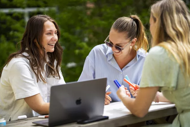 Three young females, outside at a table studying and laughing with a laptop in the middle. Photo