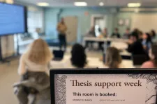 In the foreground a sign which reads "thesis support week". In the background a lecture with sutdents. Photo.